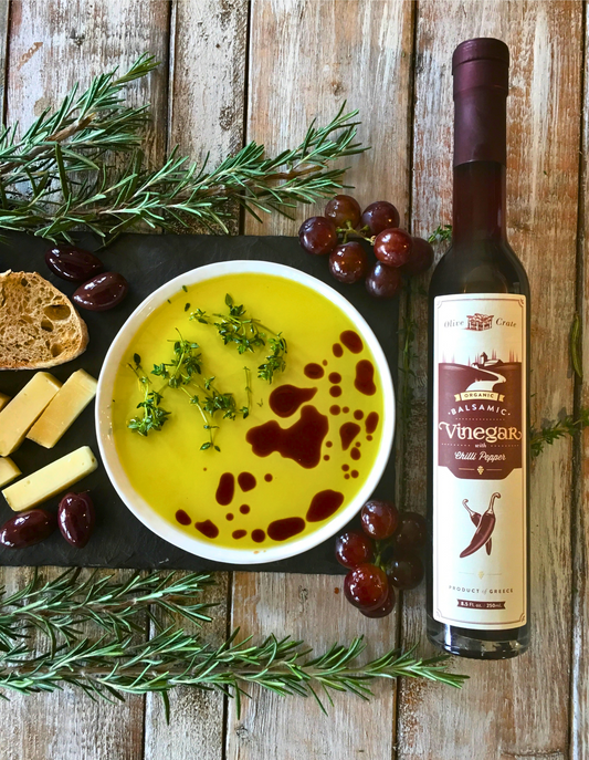 a bottle of Olive crate chili pepper balsamic next to a small bowl of Olive Crate Kores estate Olive, grapes, cheese and bread surrounded by herbs