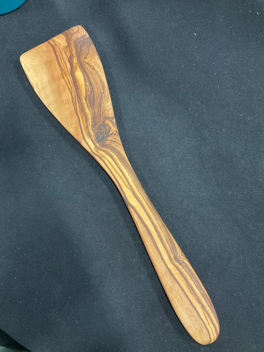 Handmade olive wood spatula on dark background, showing natural grain and warm golden tones — by Olive Crate