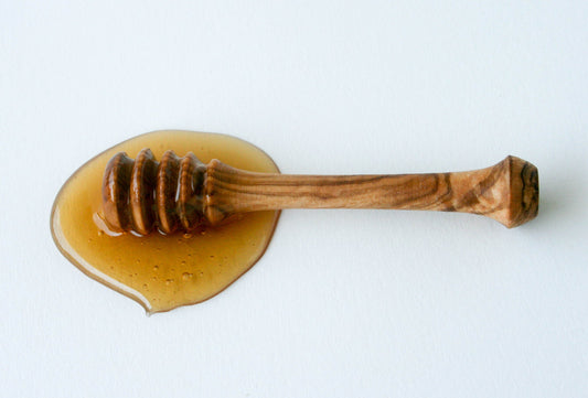 Hand-carved olive wood honey dipper resting in honey puddle, white background